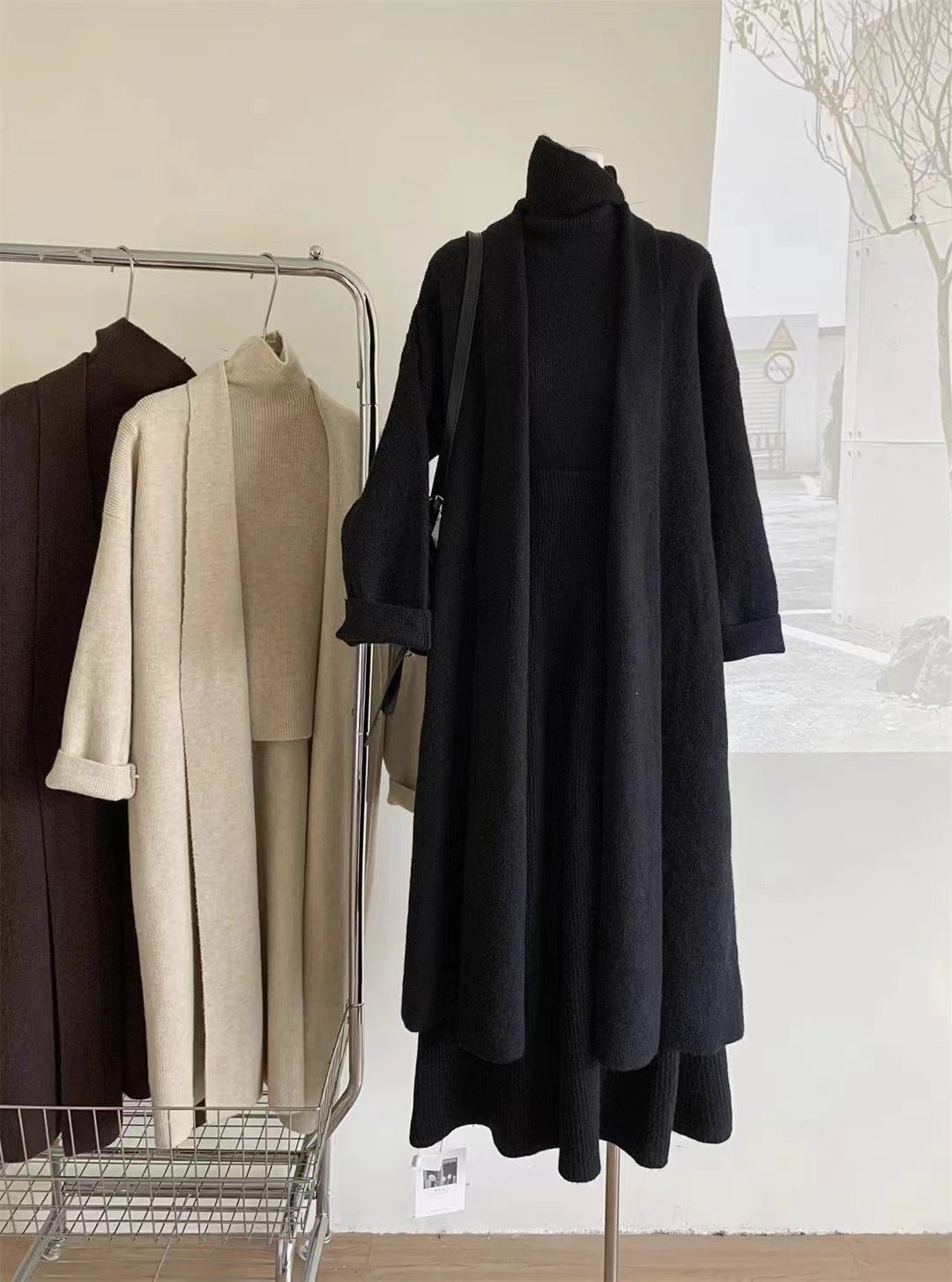 Black coat hanging on a rack with other coats against a white wall.
